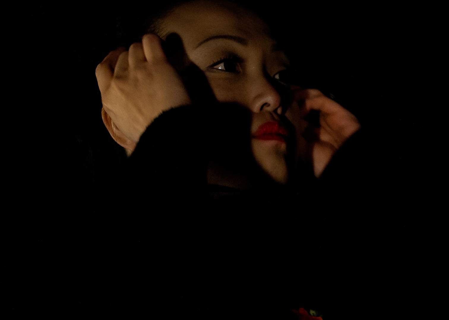 A very close up image of a woman's face, mostly cast in shadow. Her hands are tucking hair being her ears and she wears red lipstick and looks off to teh right.
